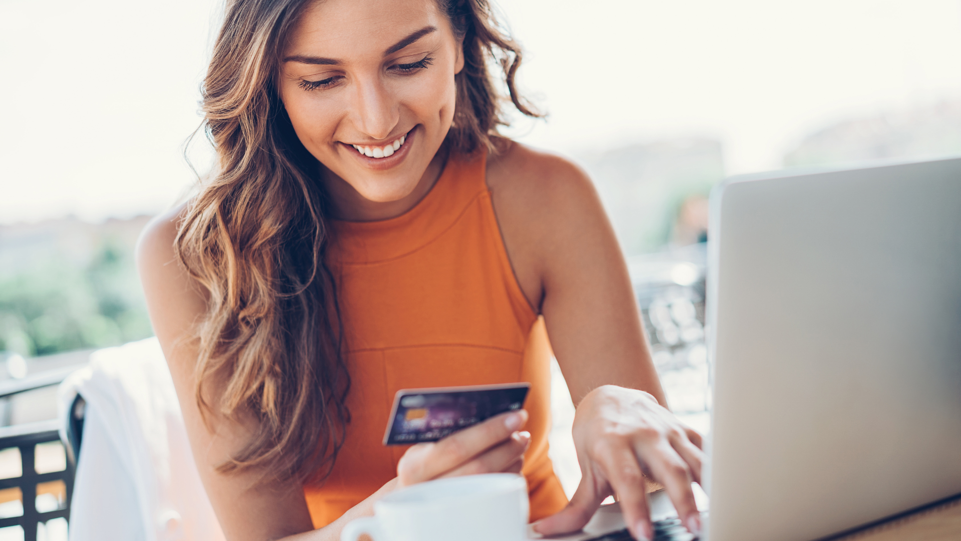 Photograph of a person wearing an orange sleeveless top holding her first credit card in one hand while using a laptop with the other hand Photograph of a person wearing an orange sleeveless top holding her first credit card in one hand while using a laptop with the other hand