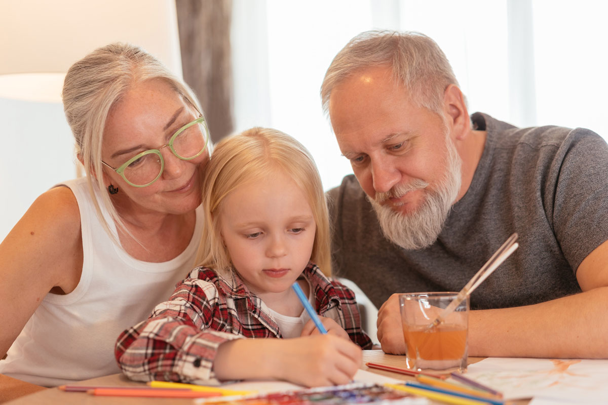 A young blonde girl is coloring with her grandparents sitting behind her