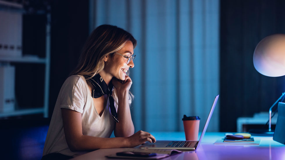 A woman is sitting in a room at night, smiling while using her laptop