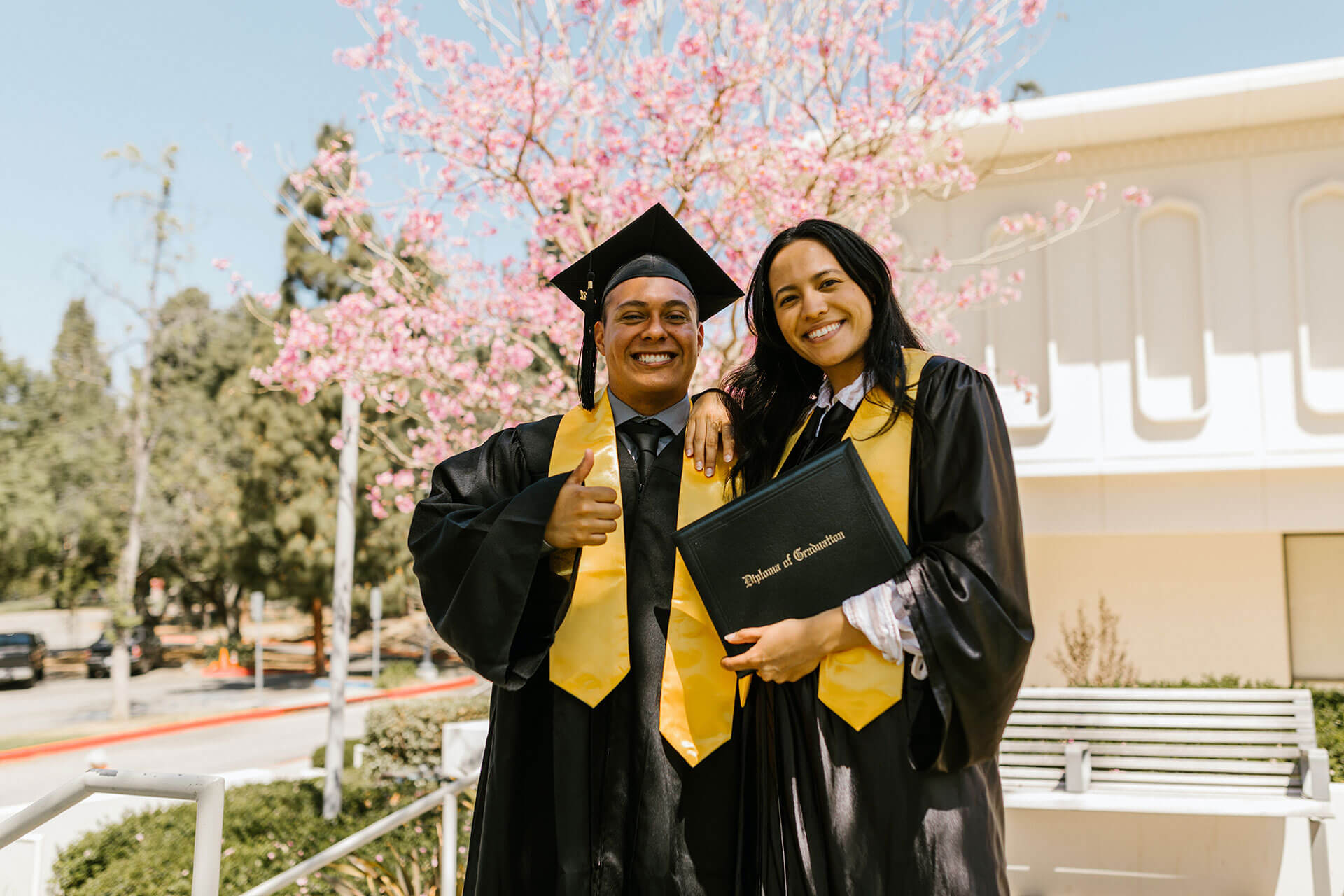 Two students are wearing a cap and gown and holding a diploma, smiling for the camera